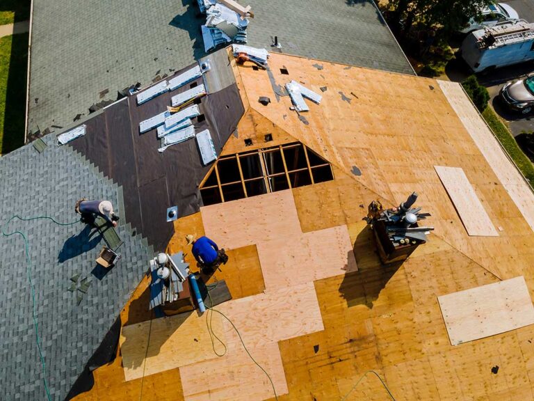 Homeowner inspecting roof shingles and cleared gutters beside a house after winter weather.